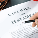 Person holding a pen over a last will and testament document on a desk.