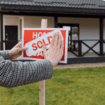 Person placing a “SOLD” sticker on a red “Home for Sale” sign in front of a house.