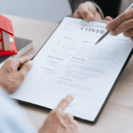 Person reviewing and signing a real estate contract with house model nearby