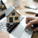 Person using laptop with small house model on keyboard and home photo on screen