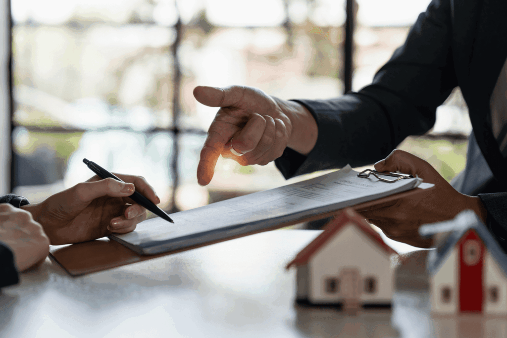 Real estate agent pointing at a contract on a clipboard while another person prepares to sign it, with small house models on the table