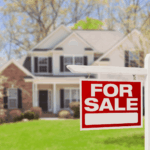 Red “For Sale” sign in front of a suburban house with green lawn and trees in the background.