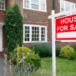 Red and white ‘House For Sale’ sign outside a brick house with a garden
