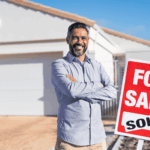 Smiling man standing with folded arms in front of a house and a red “For Sale” sign marked “Sold.”