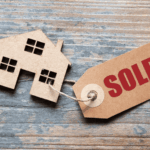 Cardboard house cutout with sold tag on wooden background.