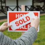Person placing a “SOLD” sticker on a red “Home for Sale” sign outdoors.