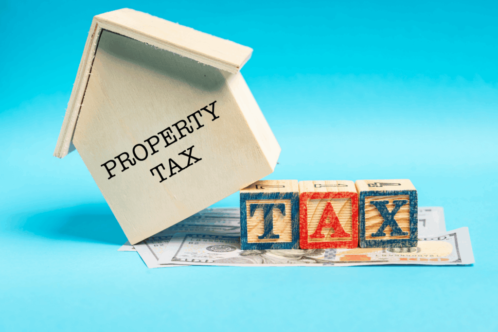 Wooden house labeled “Property Tax” beside letter blocks spelling “TAX” on top of dollar bills.