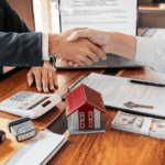 Two people shaking hands over a table with cash, contract papers, keys, and a small model house during a real estate deal.