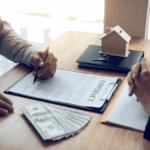 Person signing a real estate contract at a table with cash, keys, and a small house model.