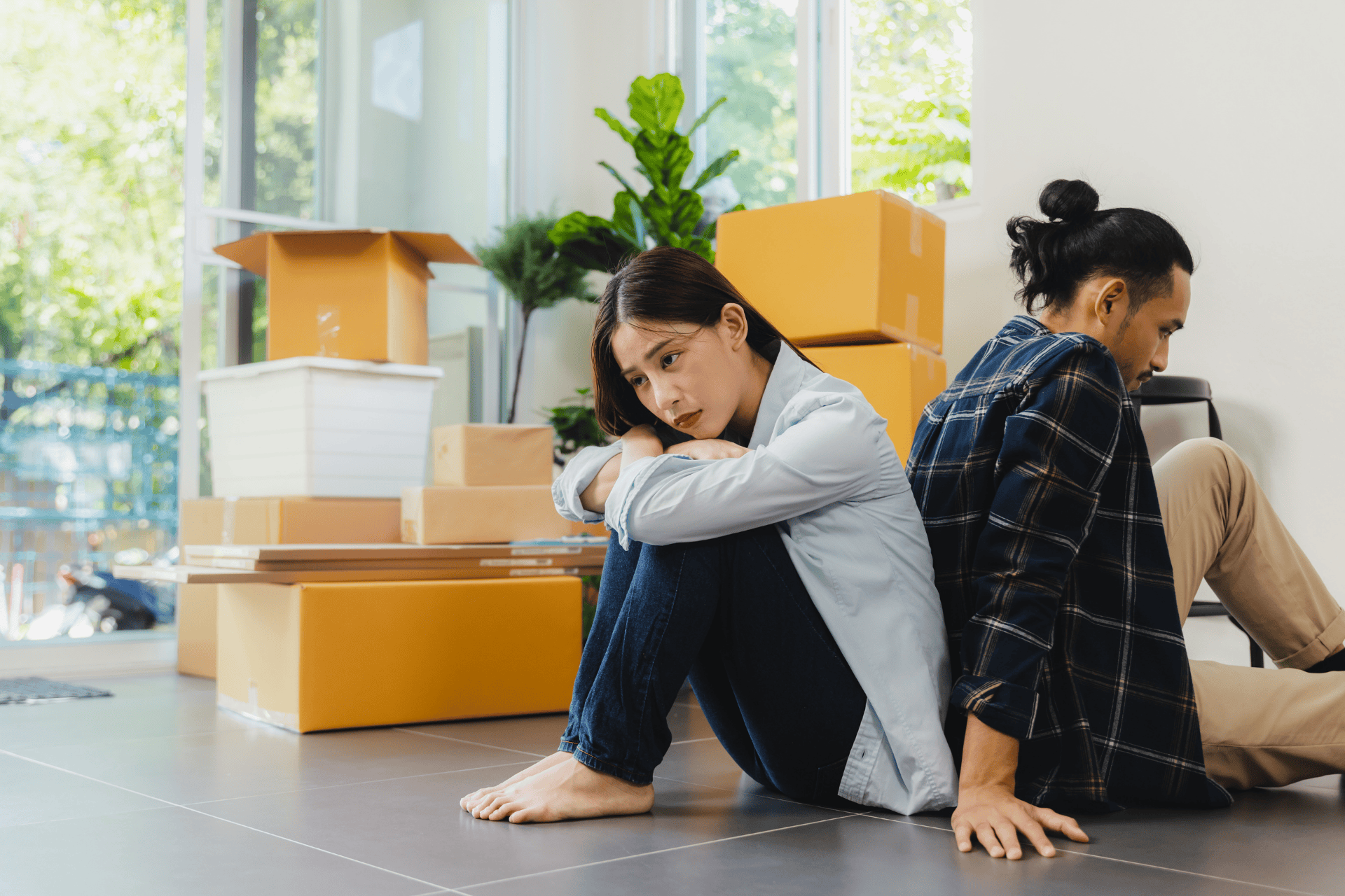 A Sad couple sitting back-to-back on the floor among moving boxes, looking upset as they go through a divorce