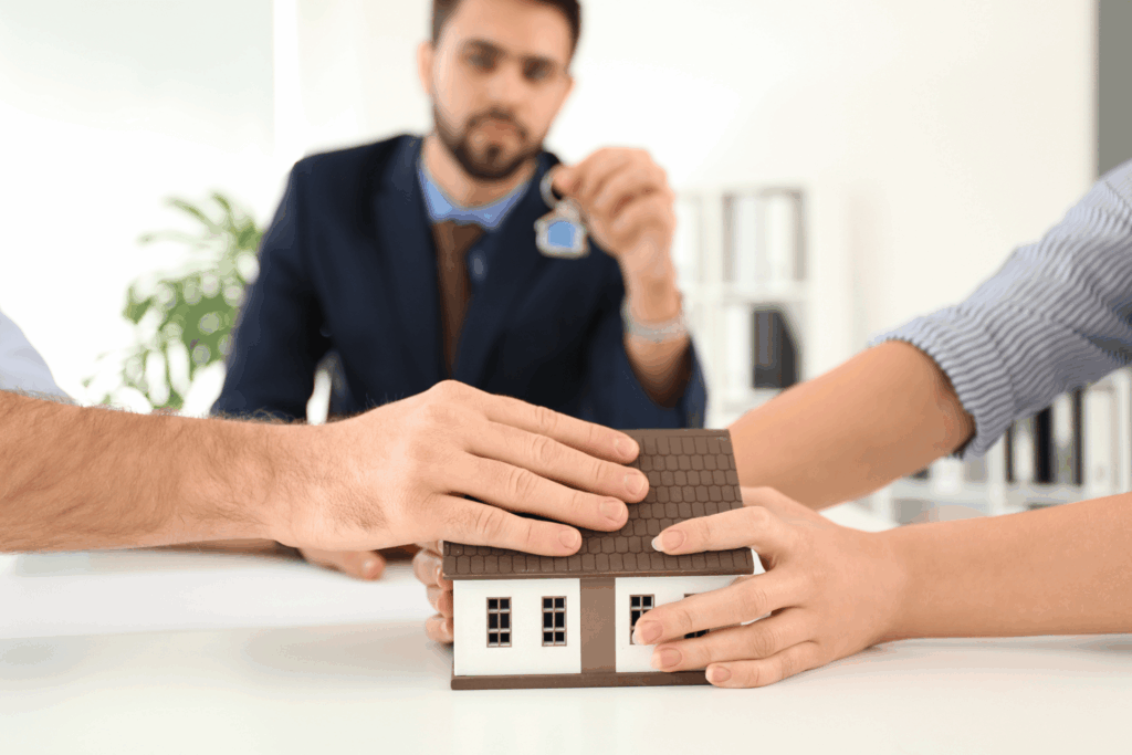 Two people hold a small model house while a realtor in the background hands over a house key.
