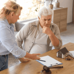 A woman shows paperwork to an older man who looks concerned, with a model house, a gavel, and scales of justice on the table.