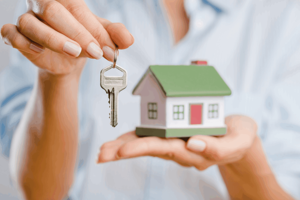 Person holding a house key and a small model home, symbolizing selling a house.