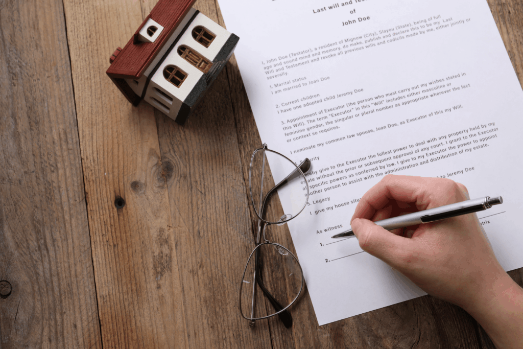 Person signing a will document next to a house model and glasses, representing a deceased estate.