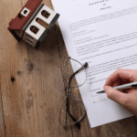 Person signing a will document next to a house model and glasses, representing a deceased estate.