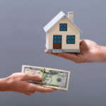 Hands exchanging cash for a small house model, representing real estate cash buyers.