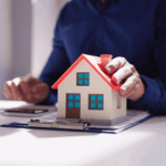 Person holding a small house model on paperwork, symbolizing estate tax considerations when selling a house.