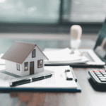 Mini house model on contract papers with pen, calculator, and laptop on desk.