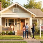Real estate agent talking to a couple in front of a beige house with a For Sale sign and moving boxes.