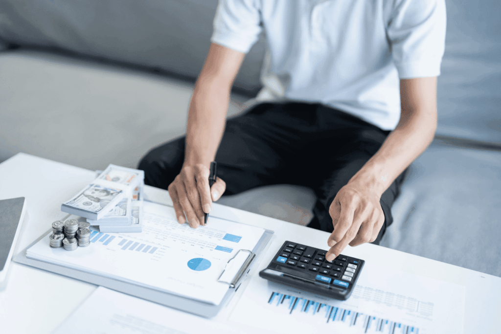 Person calculating finances with a calculator, paperwork, and stacks of cash on a desk.