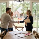 Realtor shaking hands with a couple in a bright room, with a "For Sold" sign and house visible through the window.
