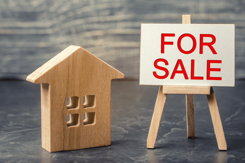 Wooden house model next to a small ‘For Sale’ sign.