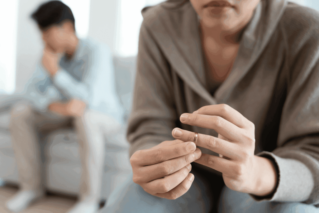 Woman holding a wedding ring in the foreground, with a distressed man sitting blurred in the background on a couch.
