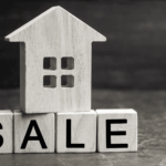 Wooden house model placed on blocks spelling “SALE,” symbolizing a home sale or real estate listing.