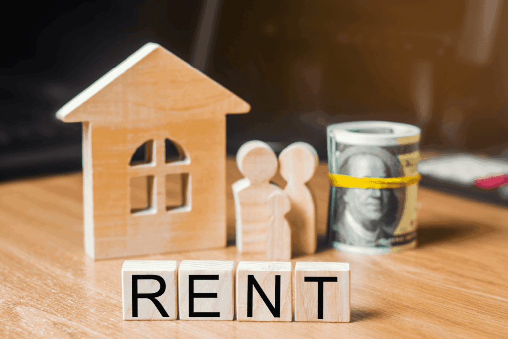 Wooden house and family figures beside rolled cash, with blocks spelling “RENT.”