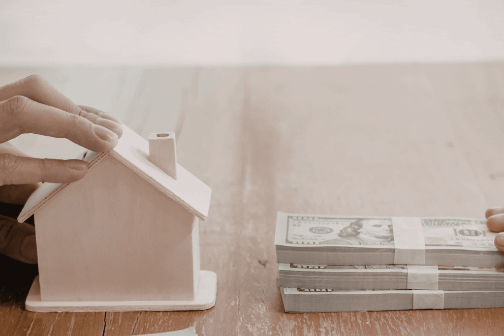 Stacks of cash being exchanged next to a small model house on a wooden table, representing the benefits of a cash offer when buying a home.