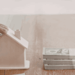 Stacks of cash being exchanged next to a small model house on a wooden table, representing the benefits of a cash offer when buying a home.