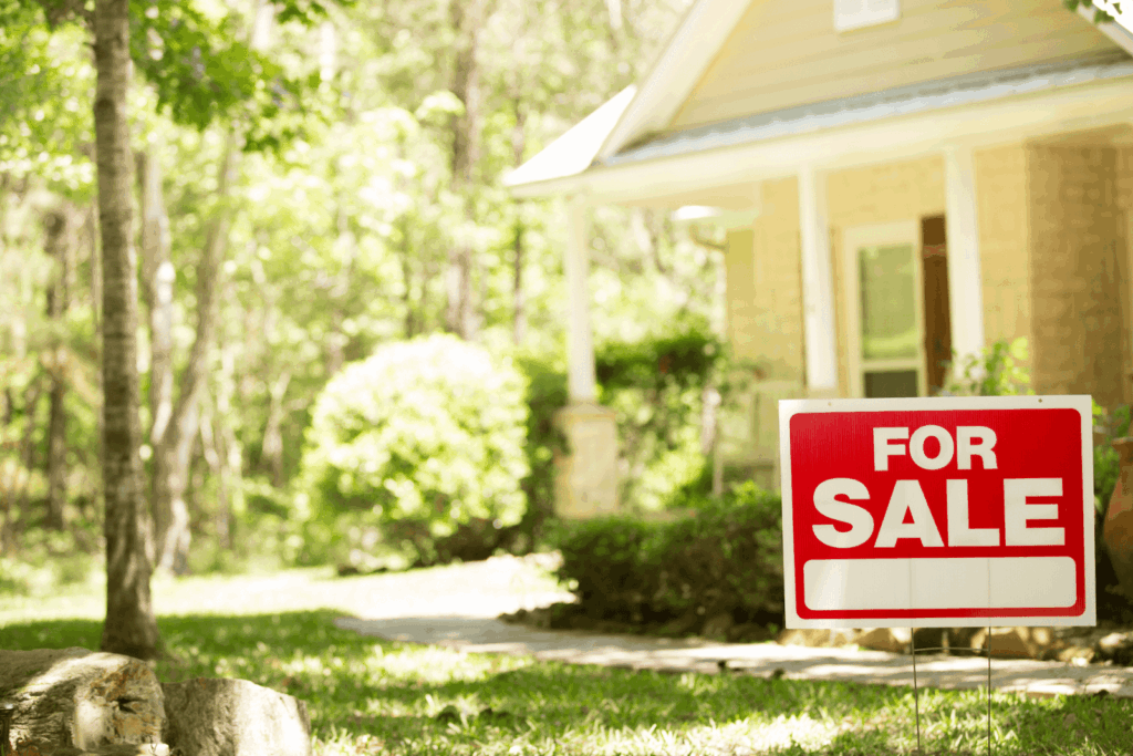 For sale sign placed in the front yard of a suburban house surrounded by trees and greenery.