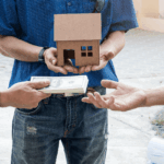 Person holding a small house model while another hand offers cash, symbolizing a property sale or real estate transaction.