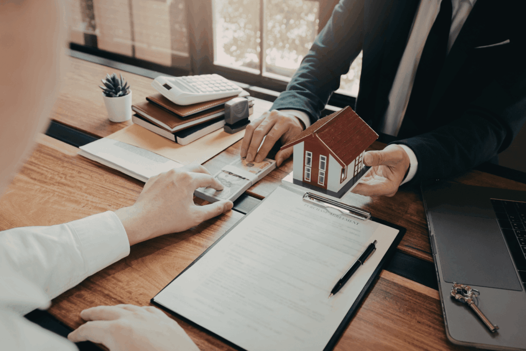 Homebuyer handing cash to a real estate agent while reviewing a purchase agreement and a small house model on a desk during a property transaction.