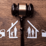 Judge’s gavel beside paper cutouts of two families with houses and cars, symbolizing property division during divorce.
