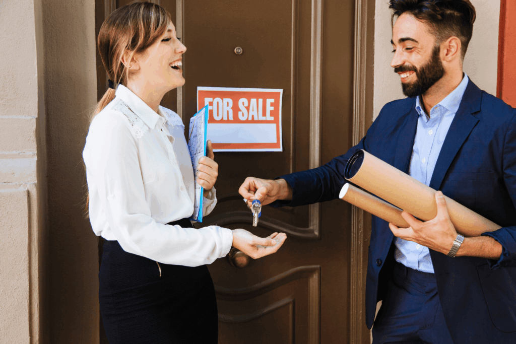 Real estate agent handing house keys to a happy homebuyer at the front door with a “For Sale” sign behind them.