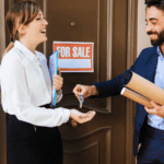 Real estate agent handing house keys to a happy homebuyer at the front door with a “For Sale” sign behind them.
