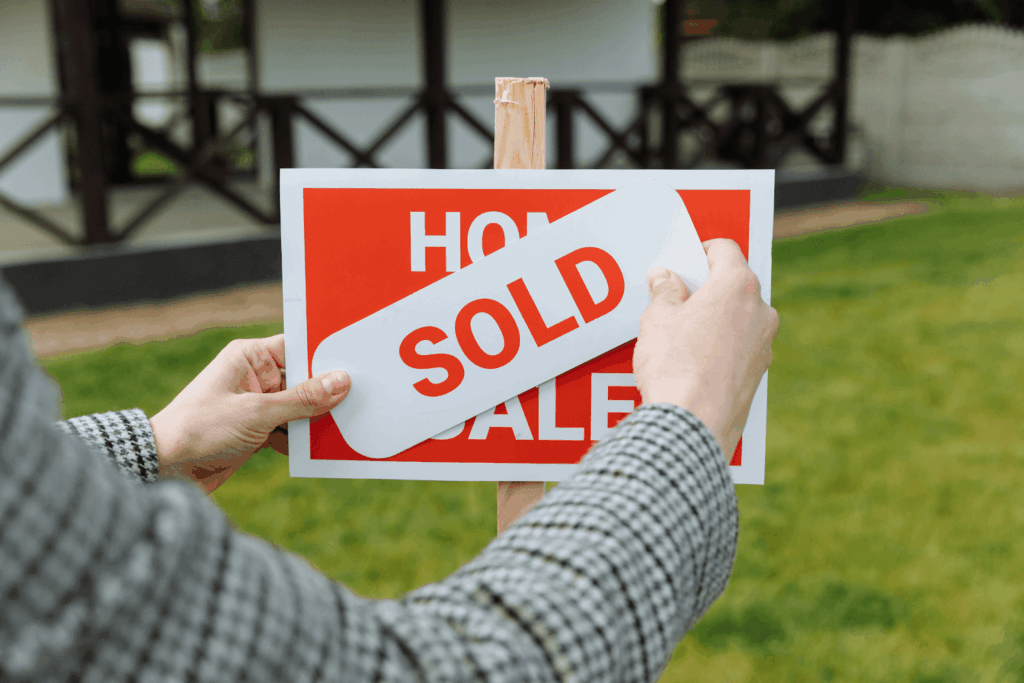 Person placing a “Sold” sticker over a “For Sale” real estate sign in front of a house, indicating a home sale completion.