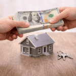 Hands exchanging a stack of cash above a small house model and keys on a wooden table, representing a quick cash home sale transaction.