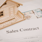 Wooden model house placed on a real estate sales contract document with architectural floor plans.