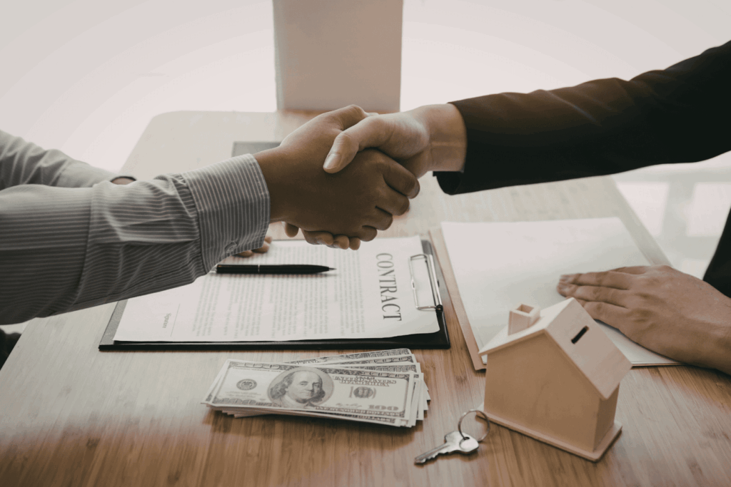  Two people shaking hands over a signed real estate contract with cash, house keys, and a small model house on the table.