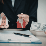 Person holding a miniature house and house keys while another hand offers cash over a real estate contract on a desk.