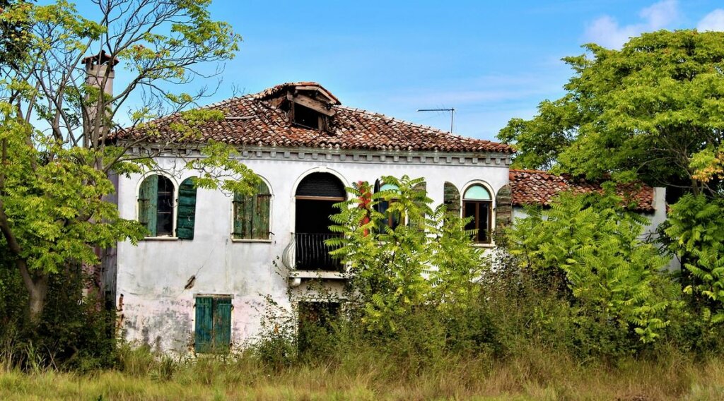 Two-story distressed house in bad shape, with broken roof and missing windows, uninhabited and looking old.