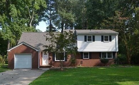 Picture of a typical two-story brick house in the Tidewater area in VA, with trees in the back, green lawn up front, with white siding covering the second floor and a one-car garage.