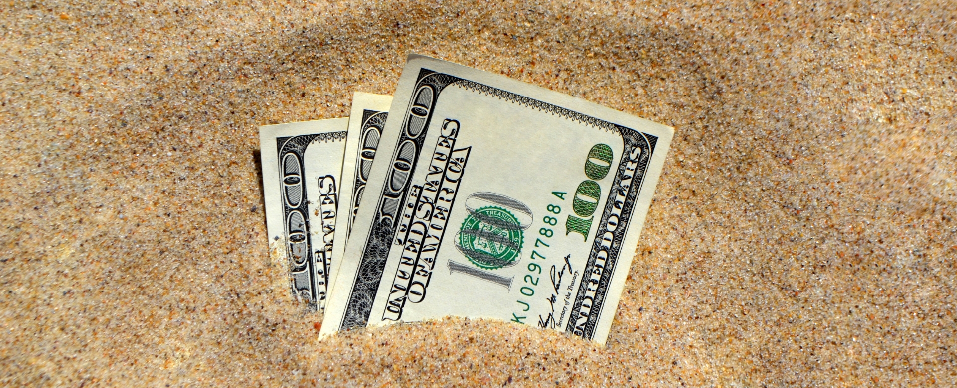 Three $100 bills sticking up out of sand.