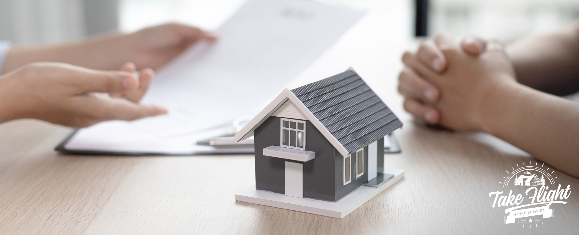 Model house on desktop with hands of two people discussing paperwork in background,