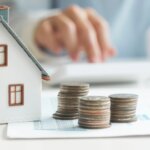 model house on desktop surrounded by stacks of coins; hands using calculator in background