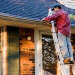 A homeowner repairing a house, cleaning gutters on a ladder to maintain the roof and improve property condition.