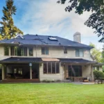 Roof repair in progress on a large home, with construction materials, ladders, and missing shingles visible against a wooded backdrop.