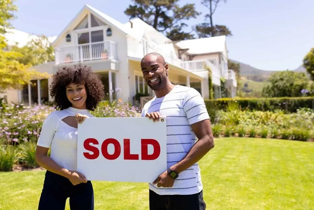 We buy houses—happy couple standing in front of their sold home holding a red "SOLD" sign on a sunny day.